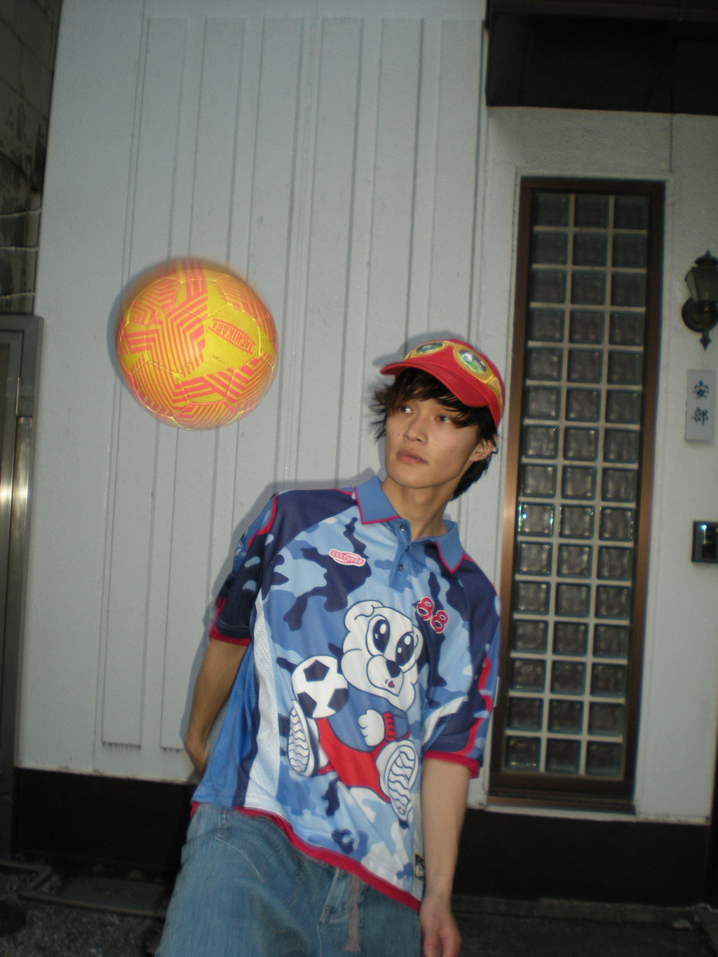 Person wearing a blue camouflage sports jersey with red accents and a cartoon design, playing ball in front of a white wall
