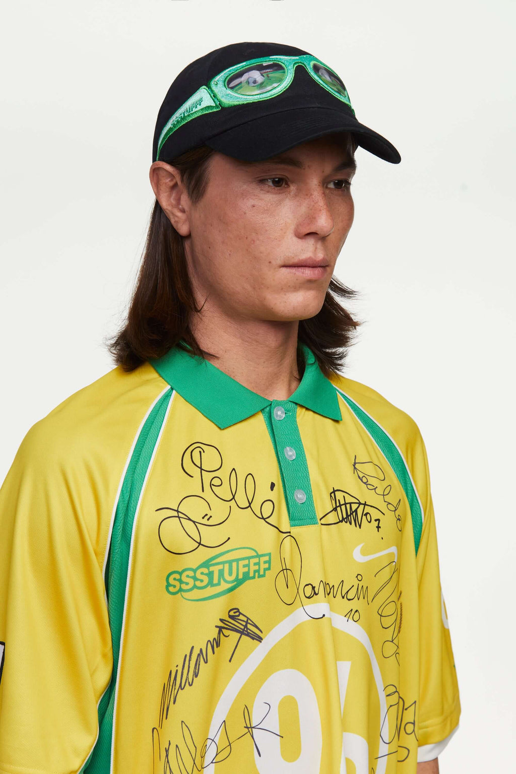 Person wearing a black cap with green sunglasses design and a yellow sports jersey with a logo, standing against a plain background. The lens of the glasses has a lenticular effect with a football ball inside a goal.