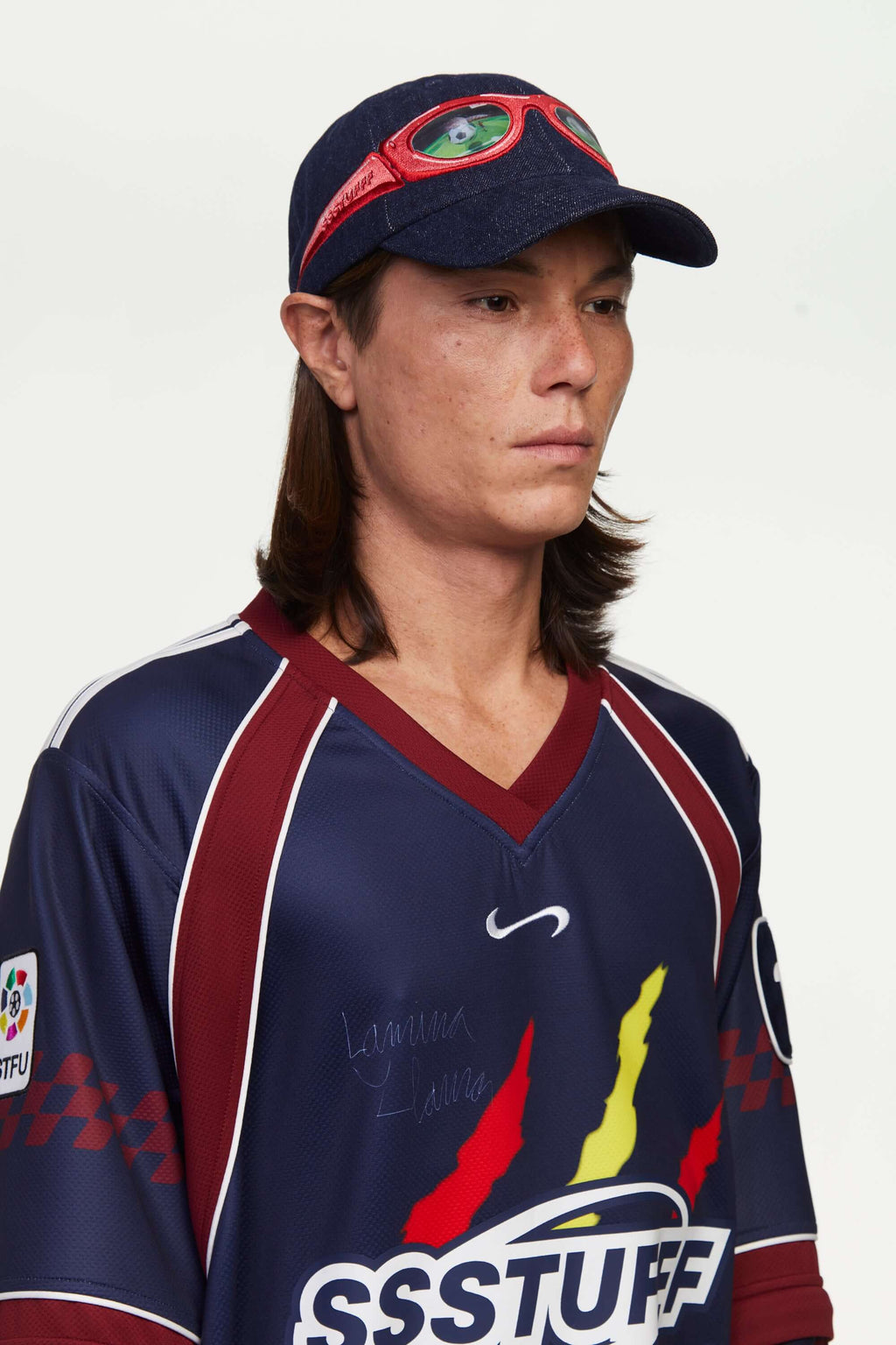 Person wearing a denim cap with red sunglasses design and a red and navy blue sports jersey with a logo, standing against a plain background. The lens of the glasses has a lenticular effect with a football ball inside a goal.