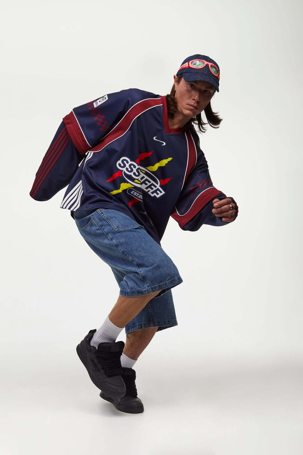 Person wearing a navy blue sports jersey with colorful graphics and text, denim shorts, and black shoes on a white background.