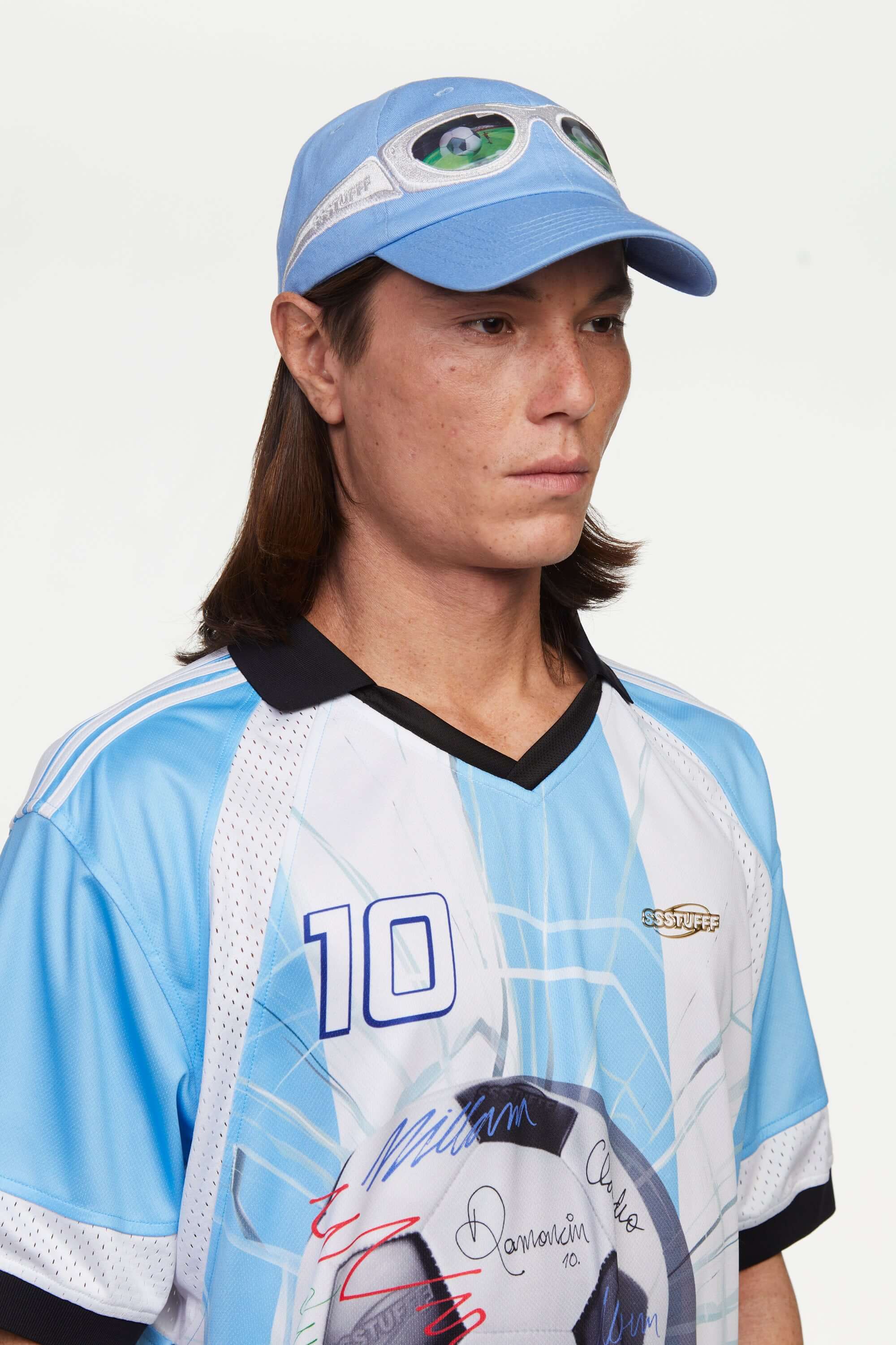 Person wearing a light blue cap with silver sunglasses design and a light blue and white sports jersey with a logo and the number '10', standing against a plain background. The lens of the glasses has a lenticular effect with a football ball inside a goal.