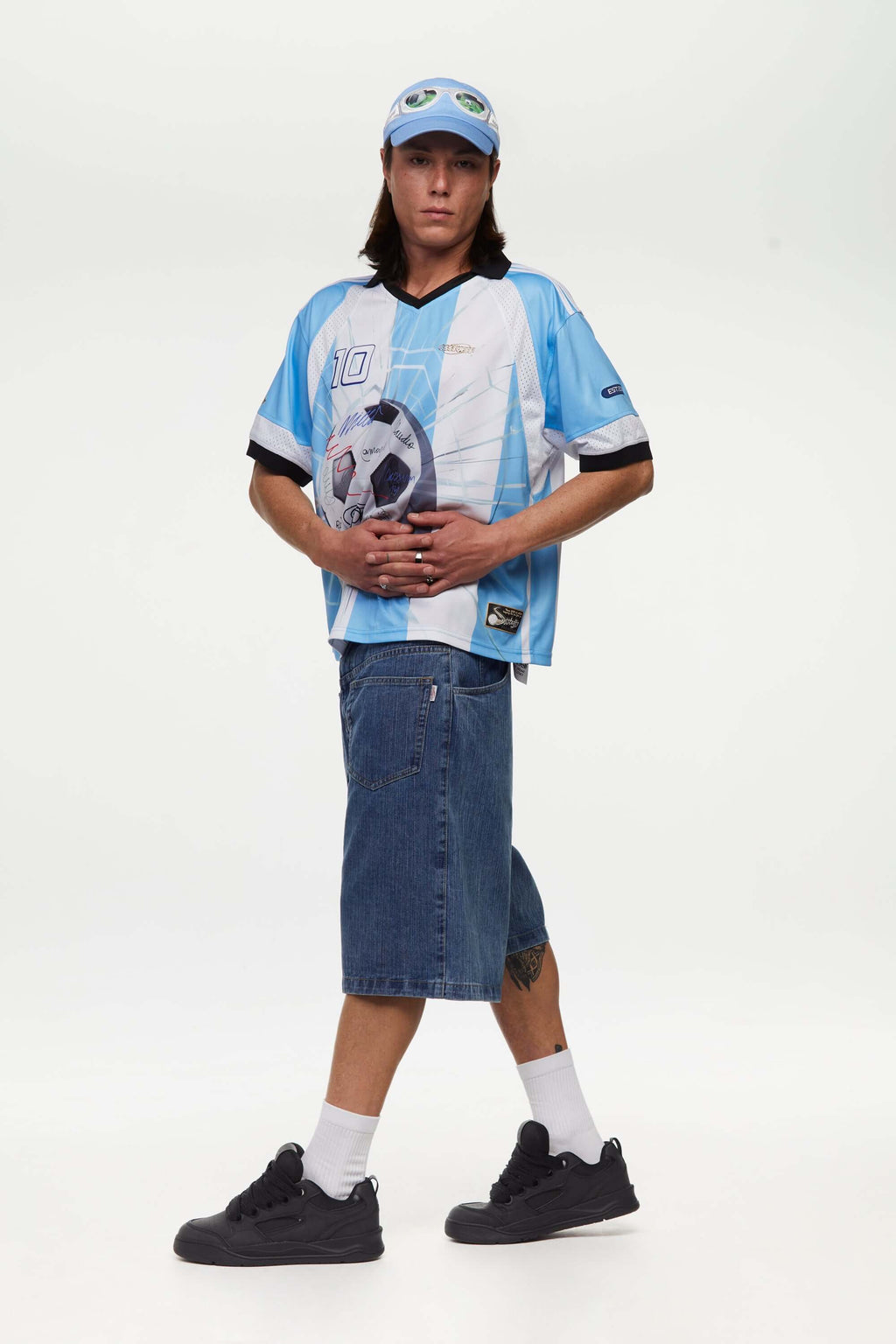 Person wearing a light blue and white sports jersey signed with a number '10' and brand logo, with a cap, denim shorts, and black shoes on a white background.