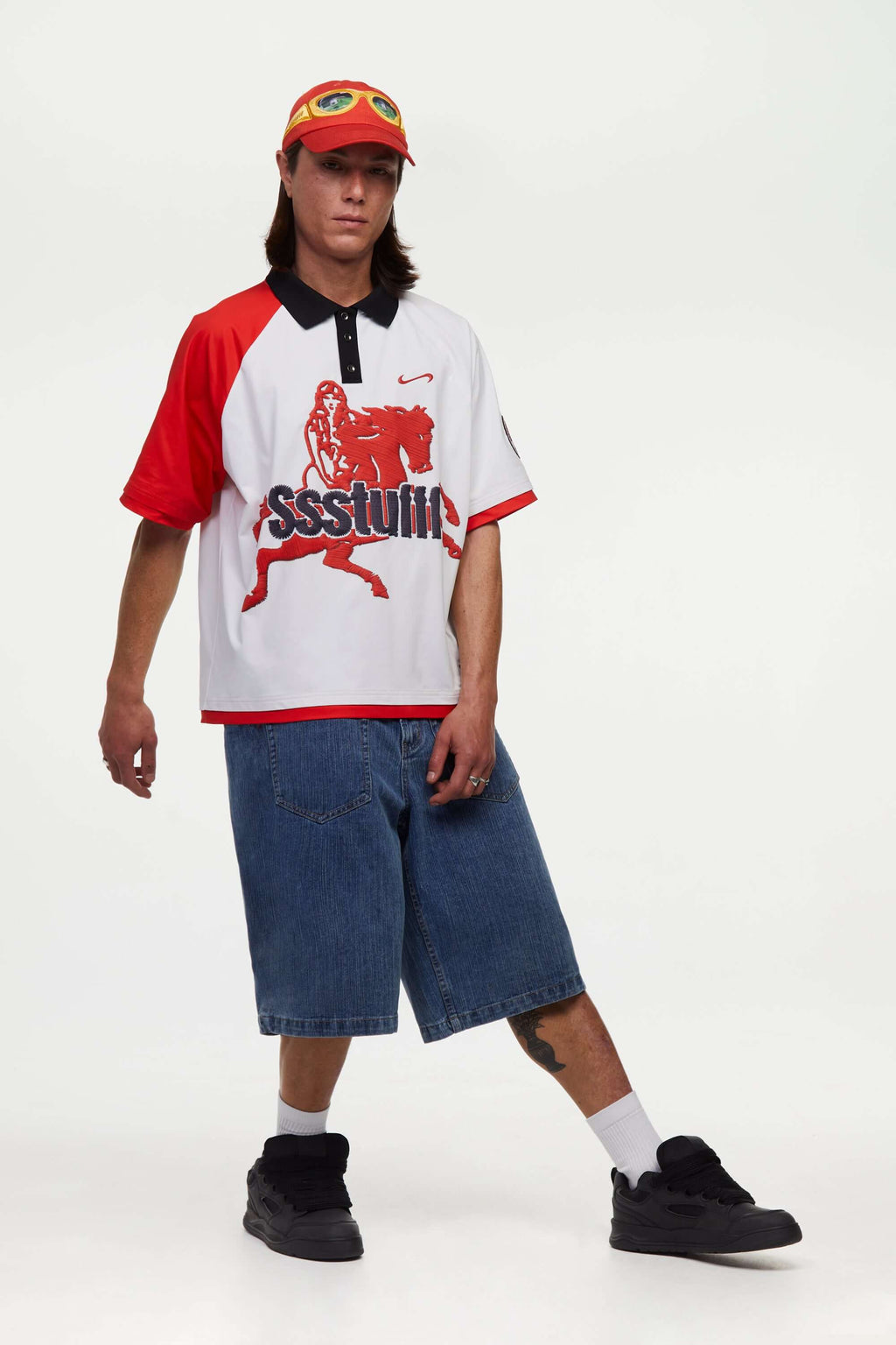 Person wearing a white football polo shirt with red accents and a print mimicking embroidered logo and motive in red and black, blue shorts, and a cap on a white background