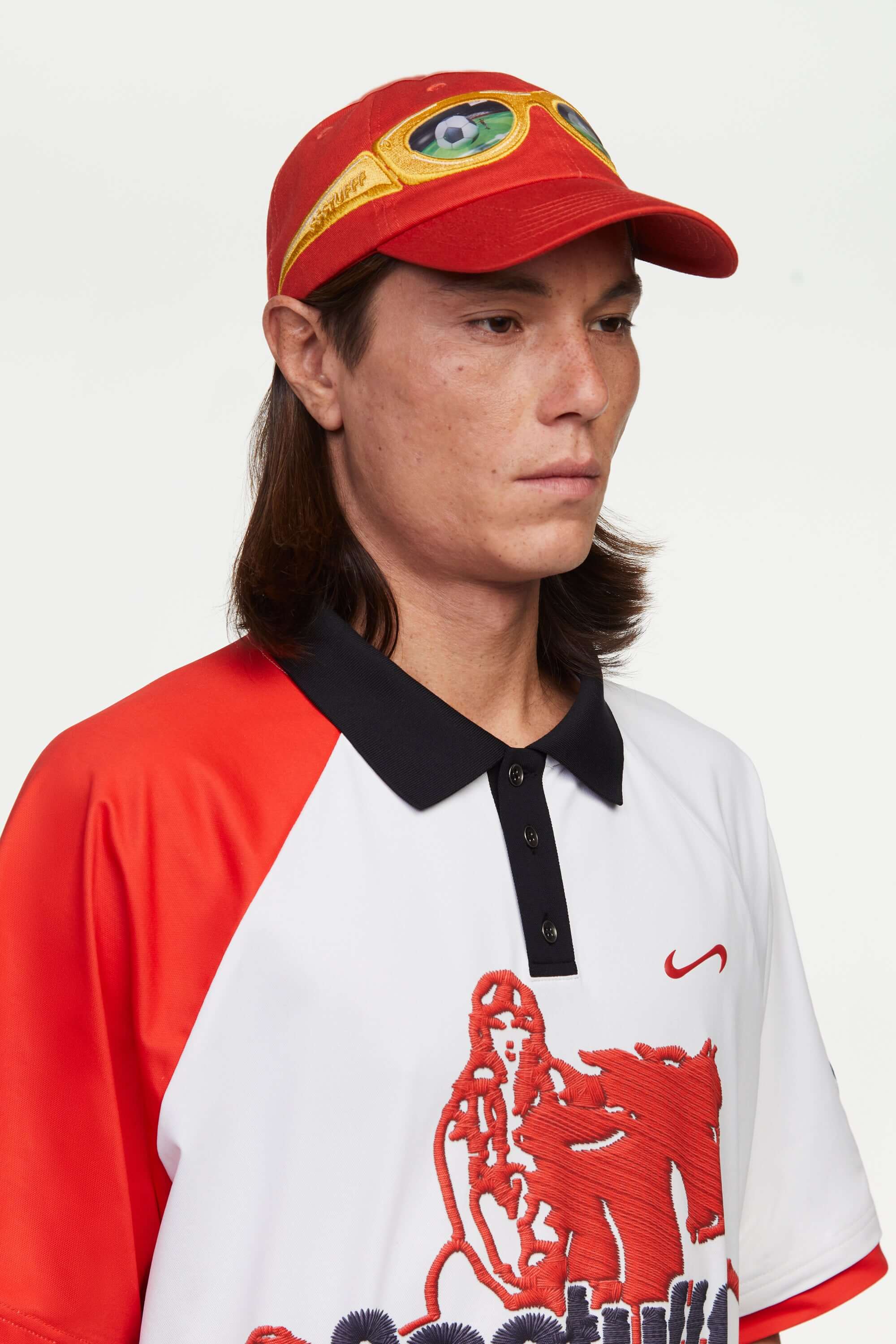 Person wearing a white football polo shirt with red accents and a print mimicking embroidered logo and motive in red and black, and a cap on a white background