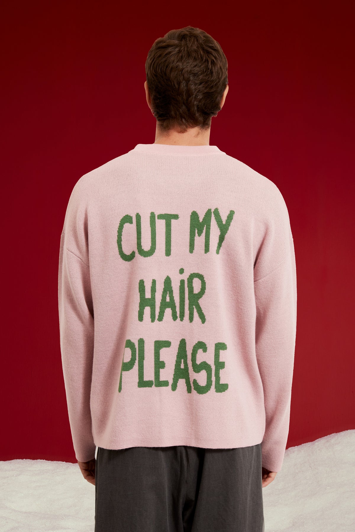 Cut My Hair Knit in pink, knitted with a character whose green fringe can be cut. Worn on a model, full-length back view, showing the CUT MY HAIR PLEASE lettering at the back.