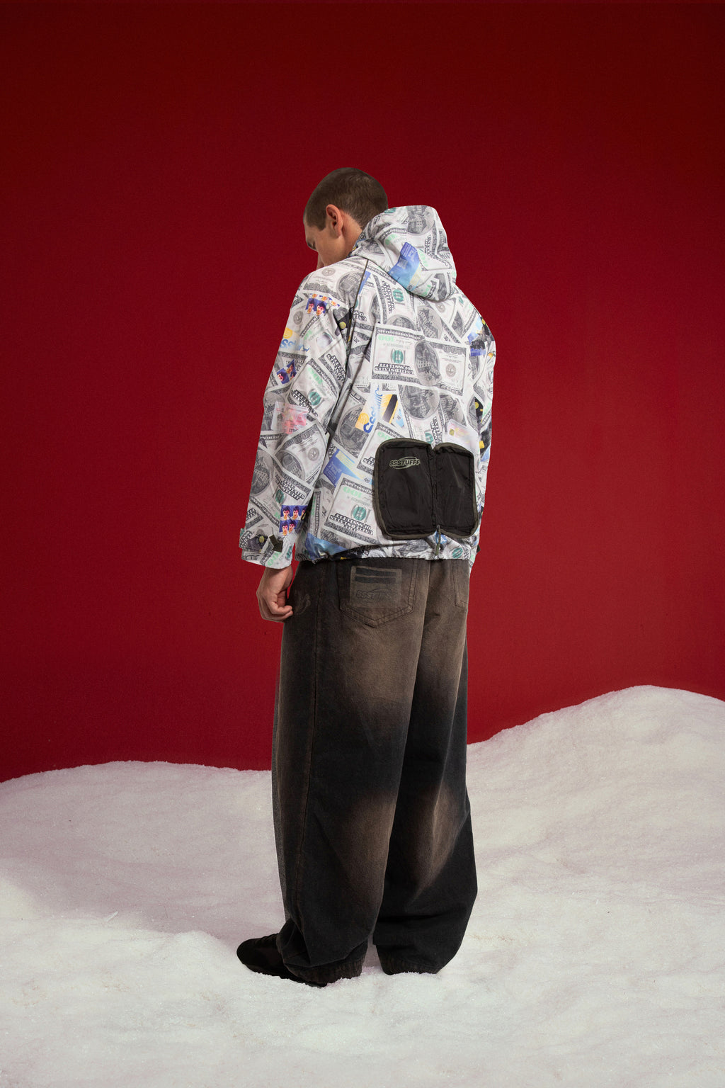 Exploding Wallet Money Windbreaker, printed all over with dollar bills and credit cards. Worn on a model, back three-quarter view, showing the black wallet pouch.