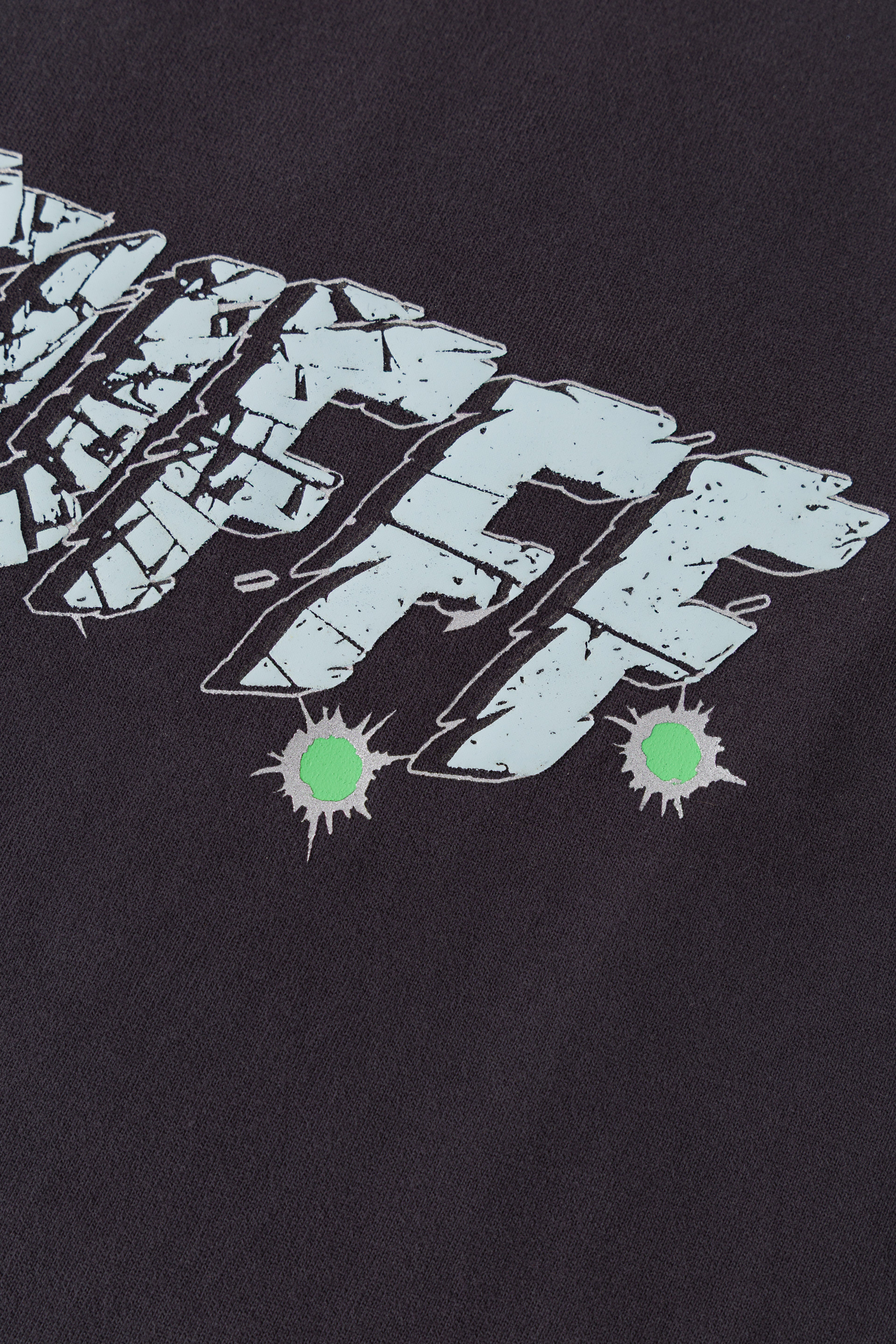 Detail of the cracked glass texture and green bullet-hole splatters on the Gunshots Tee.