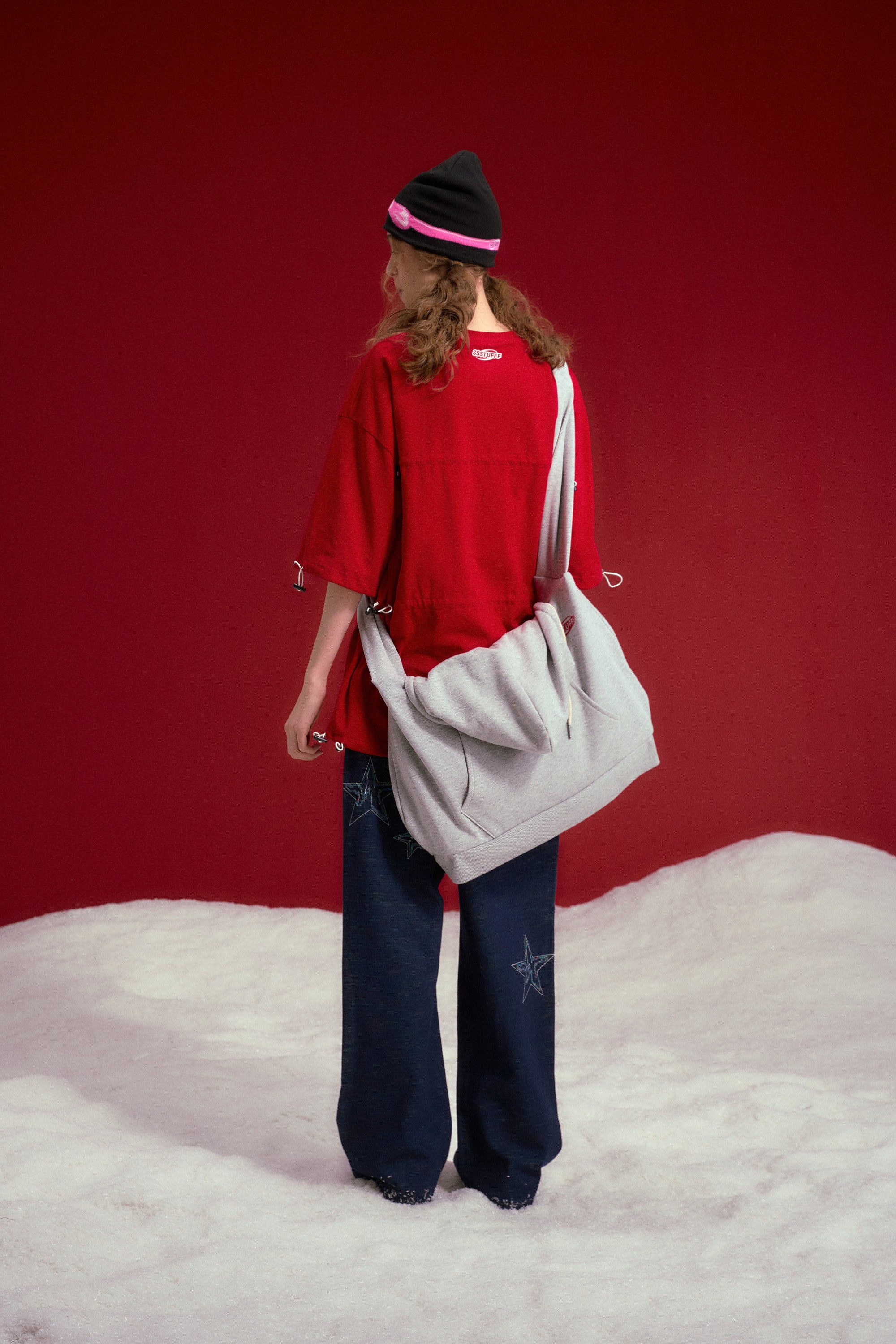 Hoodie Bag in grey marl, a hoodie rebuilt as a crossbody bag with the two sleeves knotted into the strap. Worn on a model, full-length back view.