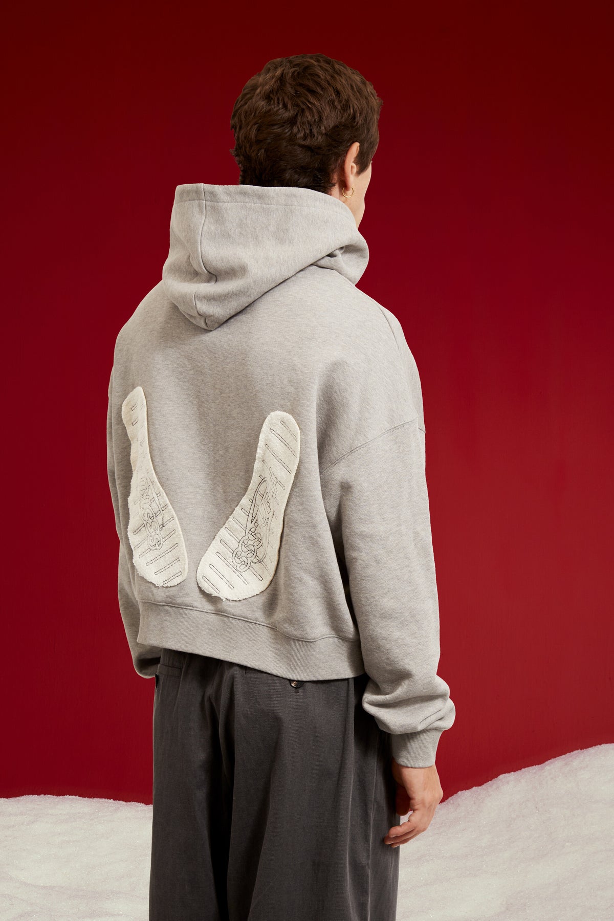 Hotel Slipers Pocket Zip Up Hoodie in grey marl, with a pair of hotel slippers tucked into the front pocket. Worn on a model, full-length back view, showing the slipper patch at the back.