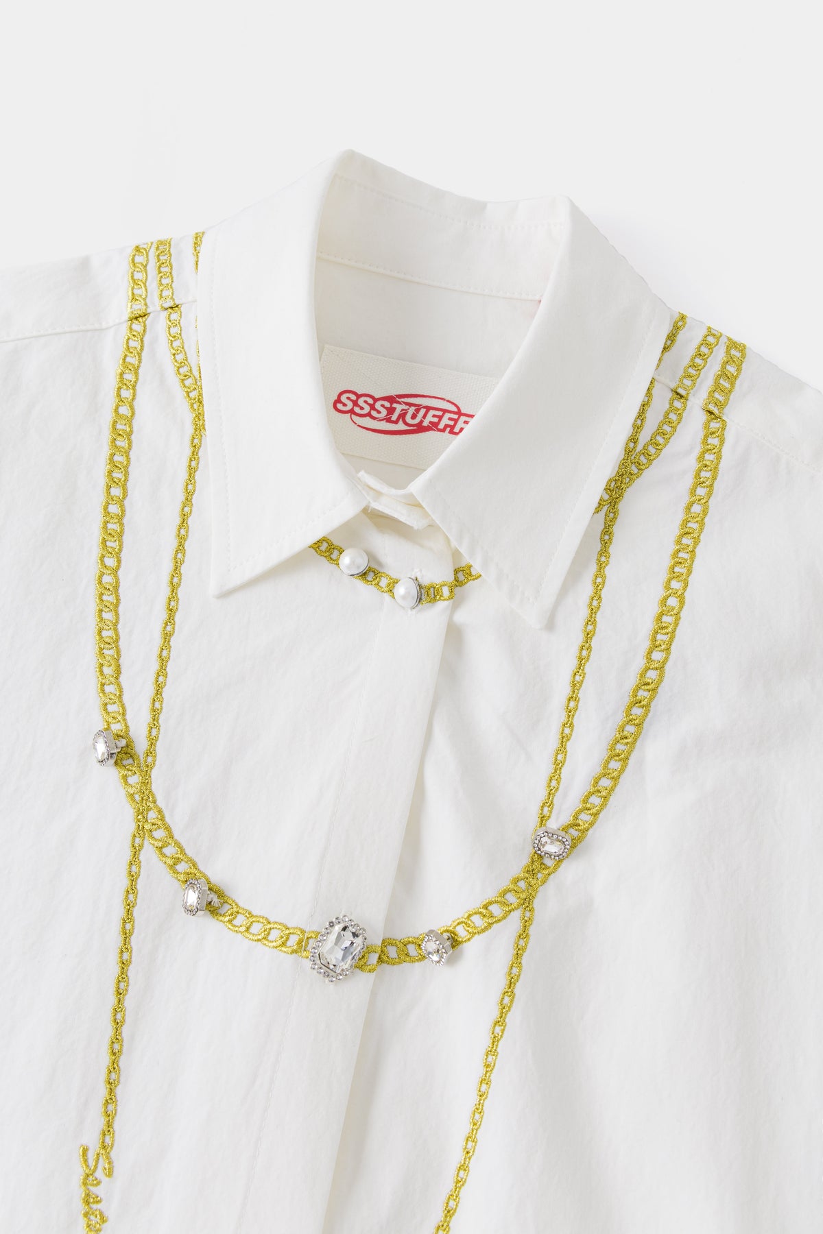 Close-up of the collar and layered embroidered gold chains, with pearls at the neck and a crystal pendant.