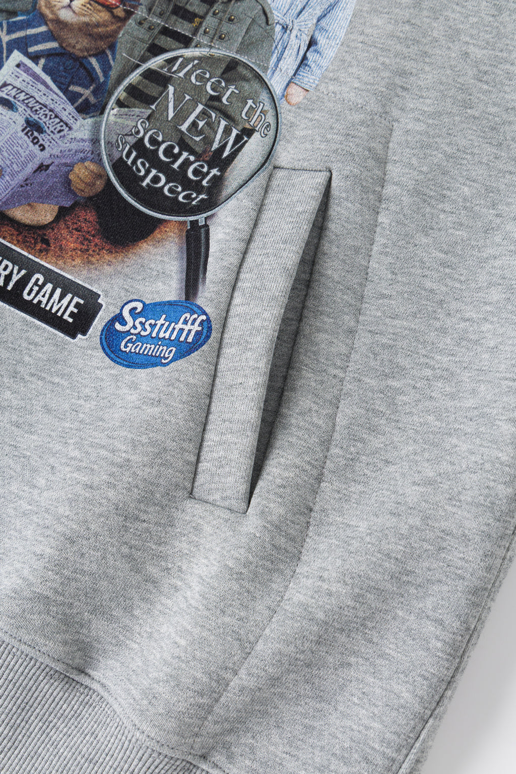Close-up of a heather gray sweatshirt featuring a graphic print with a magnifying glass, newspaper clippings, and a blue circular 'Ssstuff Gaming' logo.