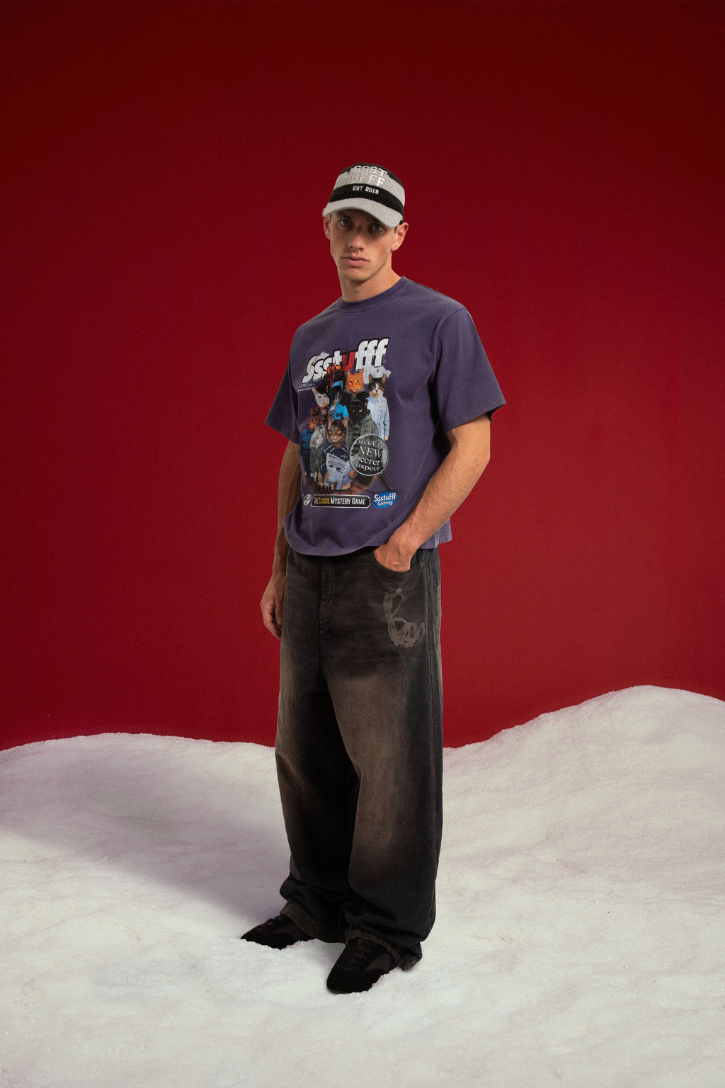 Mystery Cats Tee in washed purple, printed with the whole cast of the case redrawn as cats. Worn on a model with the Newspaper Spy Cap, full-length front view.