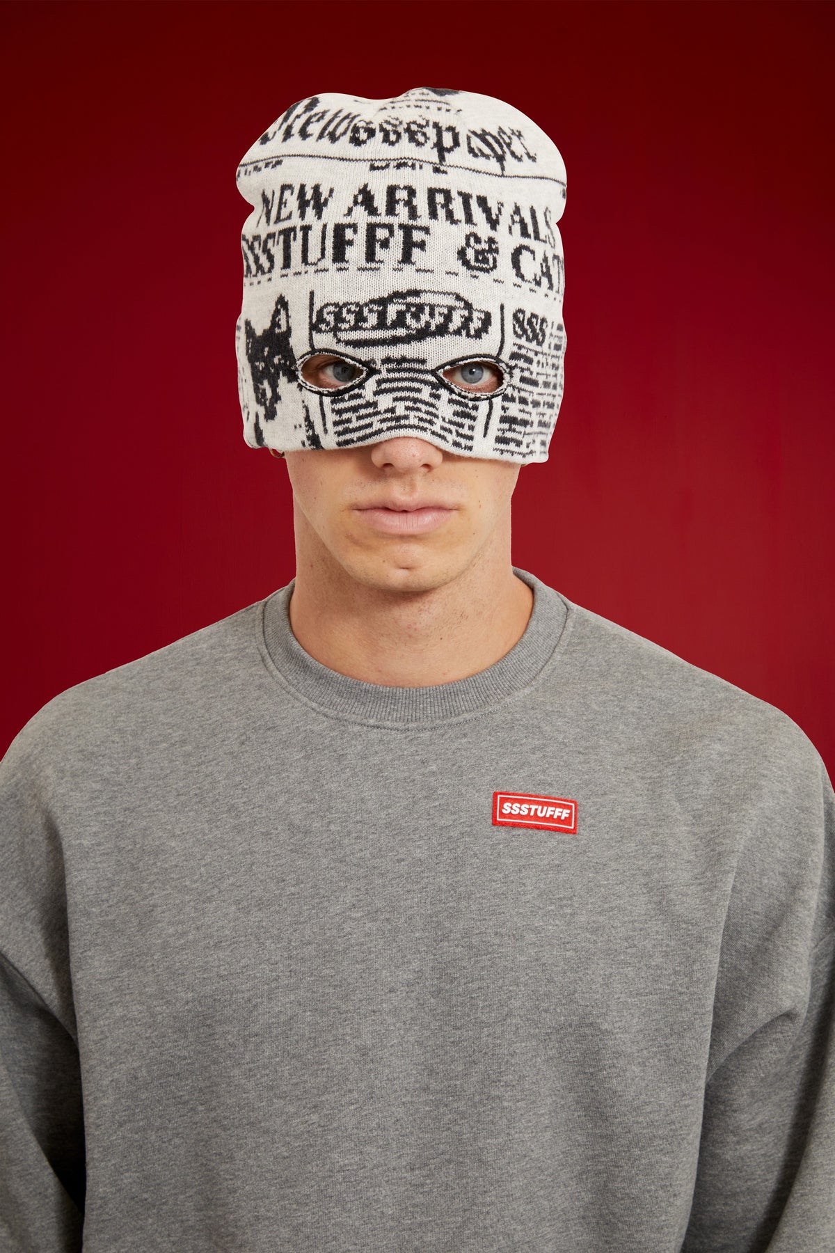 Newspaper Spy Beanie in newsprint jacquard, with two eye holes knitted into the front. Worn on a model, waist-up view, pulled down over the eyes.