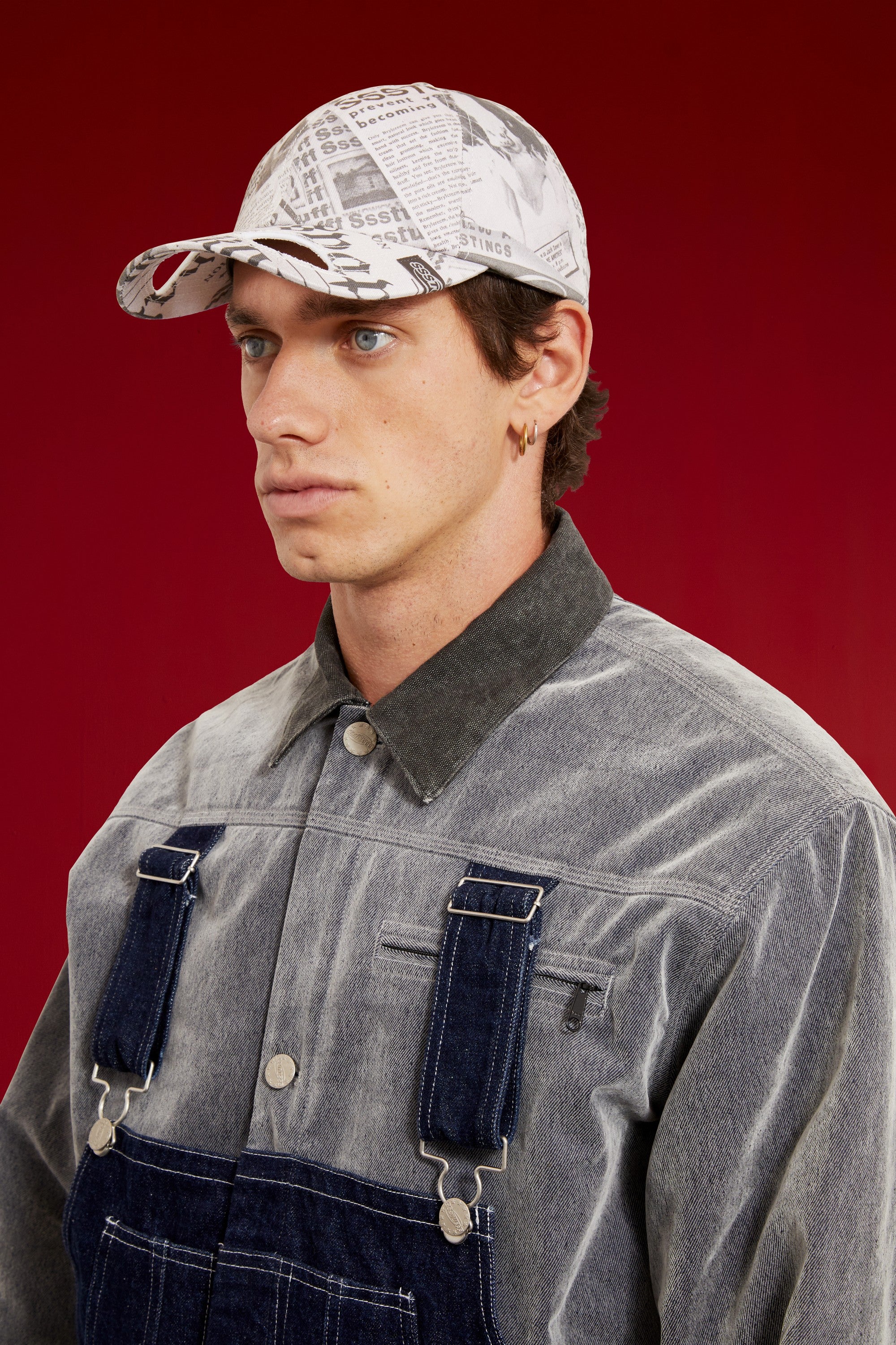Newspaper Spy Cap in all-over newsprint print, a six-panel cap with a curved brim. Worn on a model, close-up detail, front of the cap.