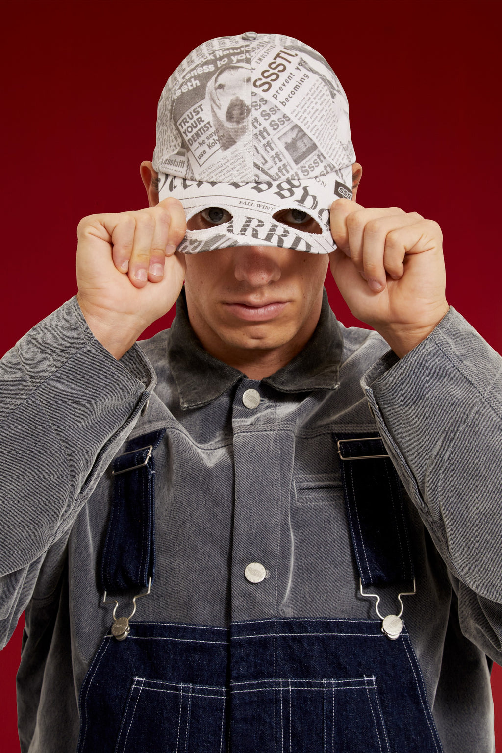 Newspaper Spy Cap in all-over newsprint print, a six-panel cap with a curved brim. Worn on a model, waist-up view, brim pulled down over the eyes.