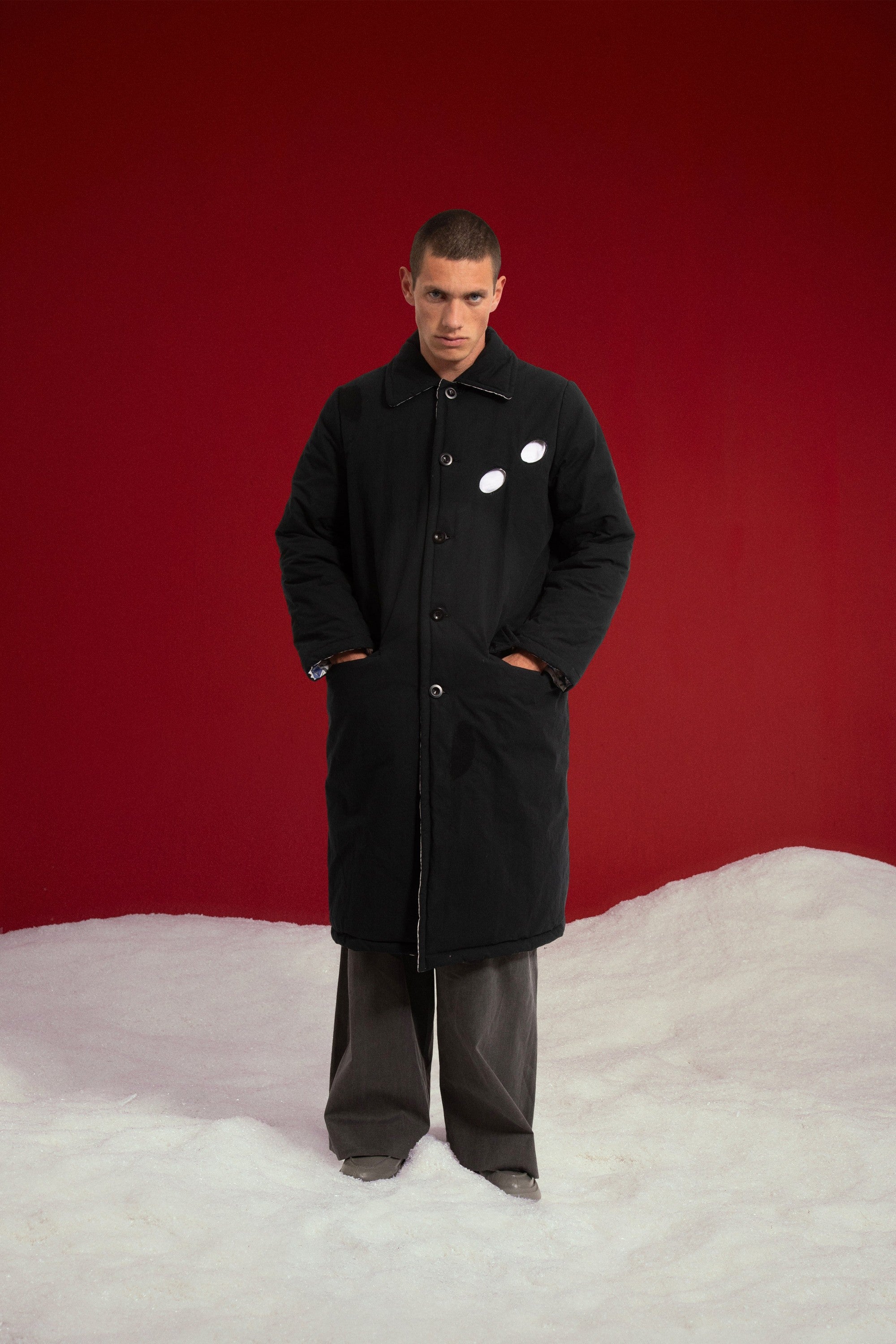 Newspaper Spy Coat reversed to its plain black side, buttoned up with hands in the pockets. Worn on a model, full-length front view.
