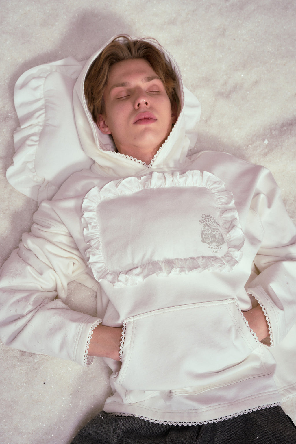 Pillow Hoodie in off-white, with a padded pillow patch sewn onto the chest. Worn on a model, waist-up view, lying in the snow.