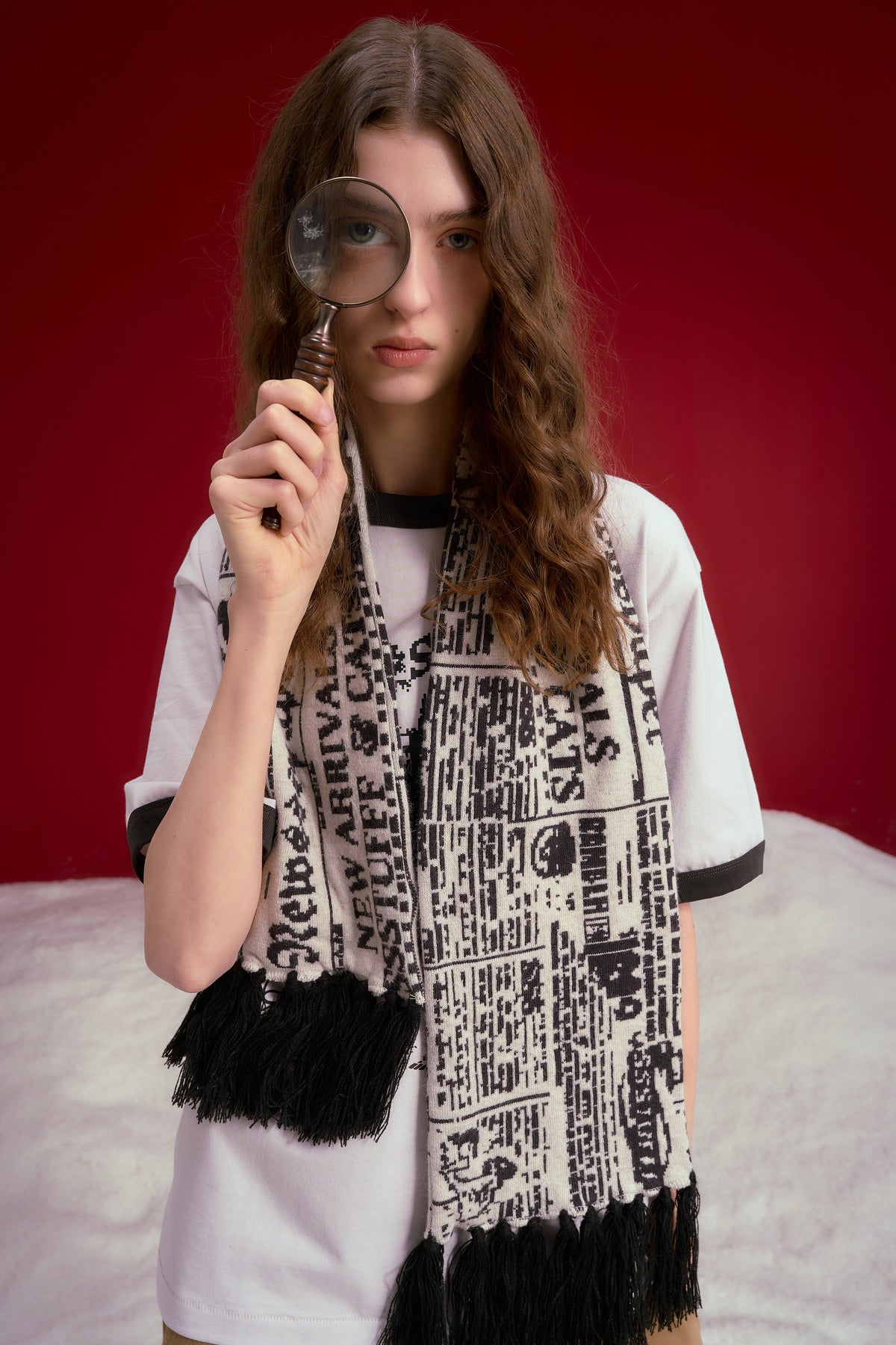 Spy Knitted Scarf in newsprint jacquard, a long scarf with black fringing at both ends. Worn on a model, waist-up view.
