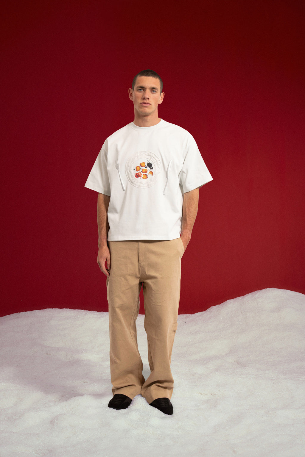 The Last Supper Tee in white, with a small plate-of-fruit print at the chest. Worn on a model, full-length front view.