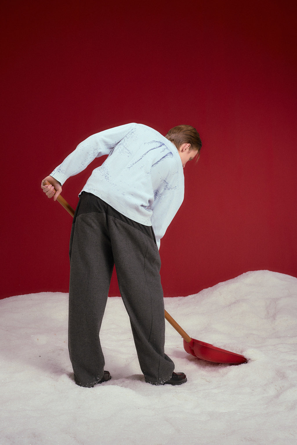 Tie Knit in pale blue, a knitted polo with the necktie knitted straight onto the front. Worn on a model, full-length back view, shovelling snow.