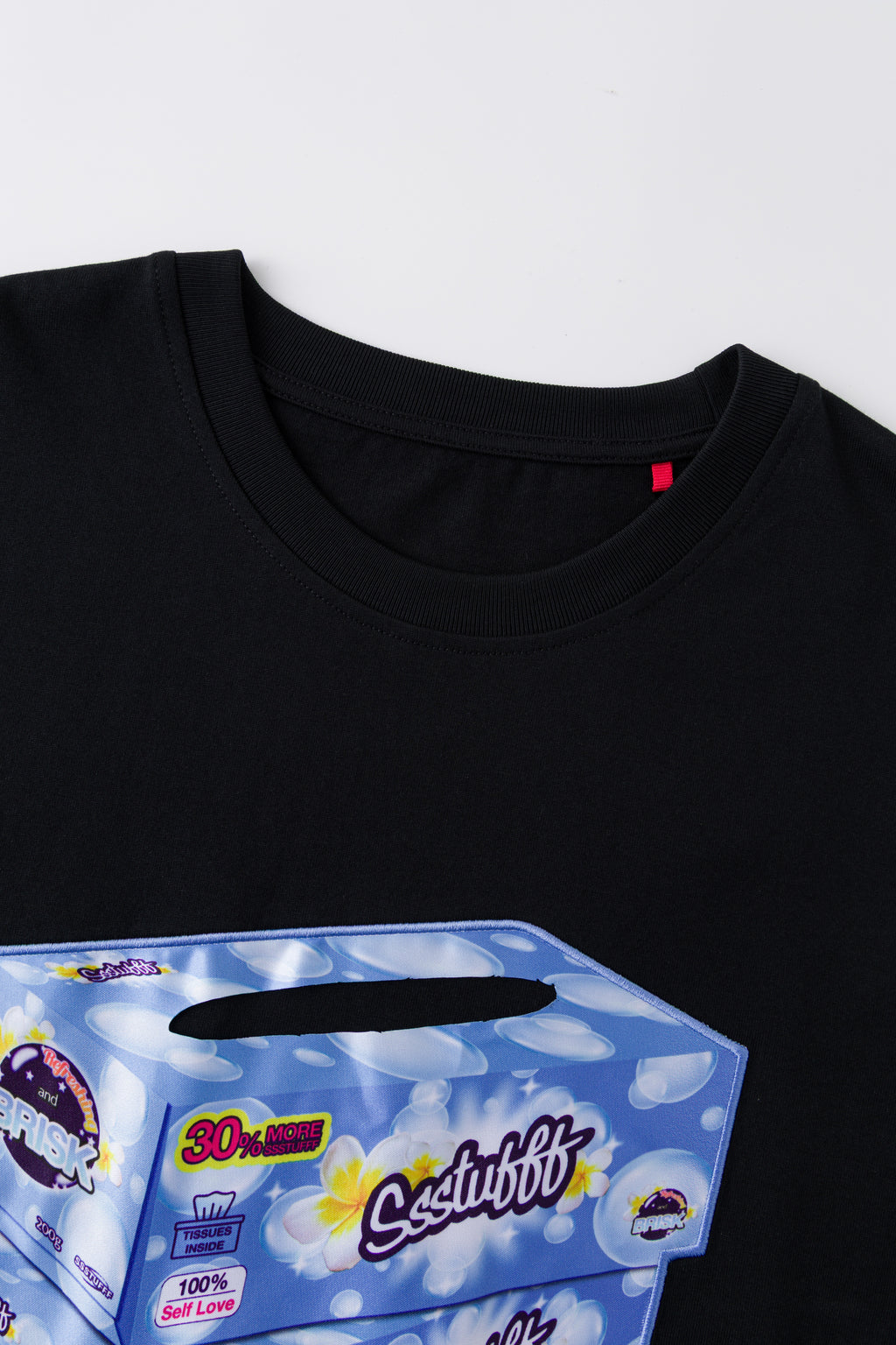 Close-up of the black tee's neckline and red woven label, above the top of the tissue box print with its slot opening.
