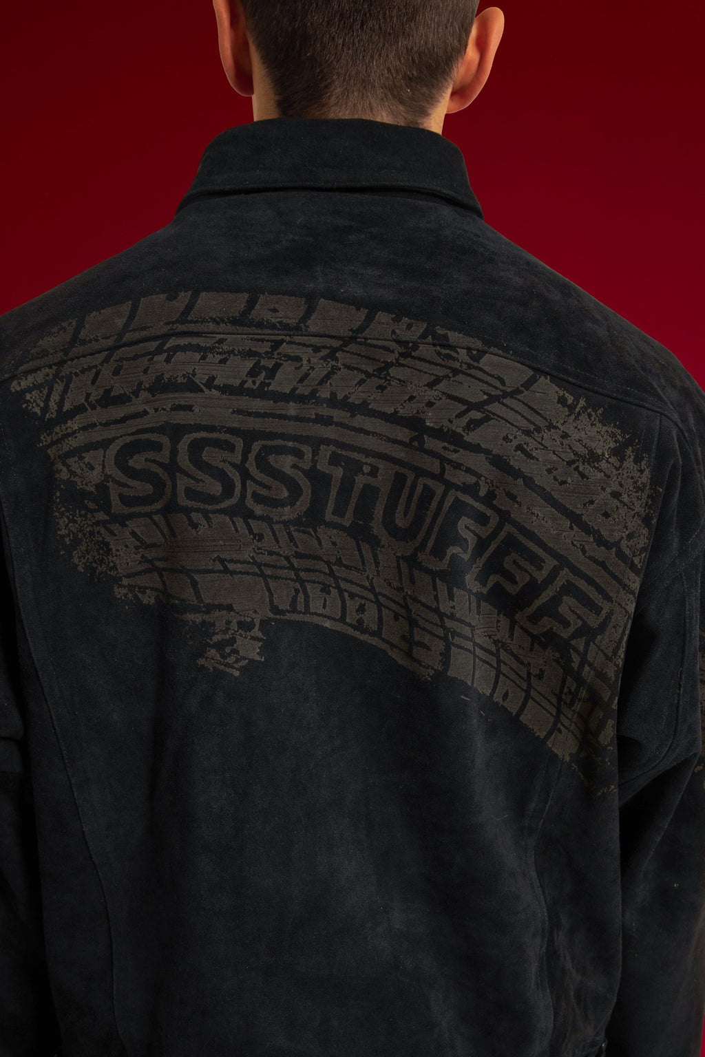 Close-up of the tyre track printed across the back of the suede jacket, SSSTUFFF set into the tread. Worn on a model.
