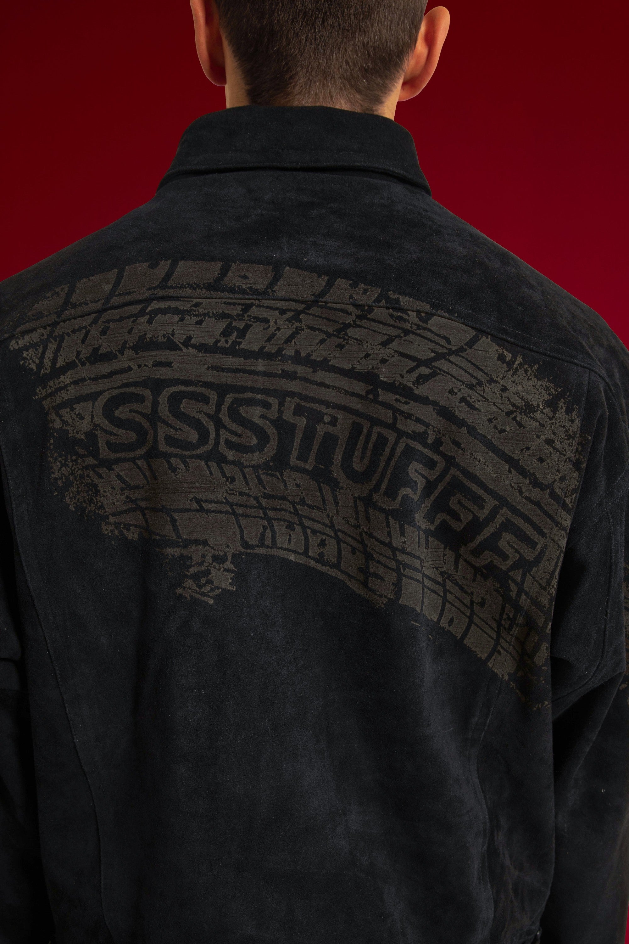 Close-up of the tyre track printed across the back of the suede jacket, SSSTUFFF set into the tread. Worn on a model.